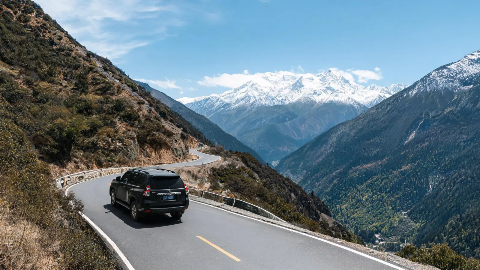An off-road vehicle driving on a winding mountain road in Western Sichuan, with distant snow-capped peaks